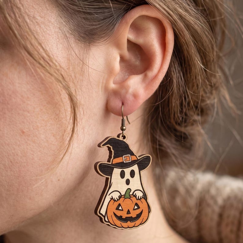 May include: A wooden dangle earring featuring a light-colored ghost wearing a black witch hat with an orange band. The ghost is holding an orange jack-o'-lantern with a black carved face. The earring hangs from a simple metal hook.