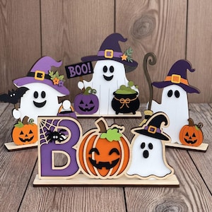 Könnte beinhalten: Ein Set aus hölzernen Halloween-Tischdekorationen mit weißen Geistern in violetten Hexenhüten, orangefarbenen Kürbissen, einem schwarzen Kessel, einer Spinne und einem Schild mit der Aufschrift BOO, montiert auf flachen Holzsockeln.