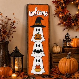 Peut inclure: Un panneau rectangulaire en bois orange avec le mot welcome écrit en blanc en haut. Trois fantômes blancs sont empilés verticalement en dessous. Le fantôme du haut porte un chapeau de sorcière noir et tient une citrouille, celui du milieu tient une chauve-souris noire et celui du bas tient un bonbon en forme de maïs.