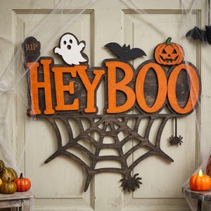 May include: A wooden wall sign featuring the orange text HEYBOO with a white ghost, a black bat, and an orange jack-o'-lantern on top. The sign includes a dark brown spiderweb cutout at the bottom with two small black spiders hanging from it.