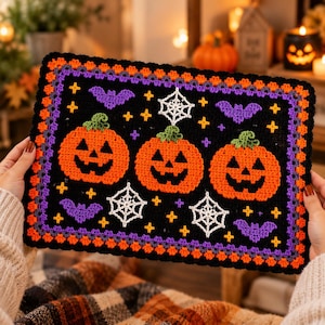 Peut inclure: Une tenture murale rectangulaire en crochet sur fond noir avec trois citrouilles d'Halloween orange, des chauves-souris violettes et des toiles d'araignée blanches. La bordure présente des motifs en crochet orange et violet.