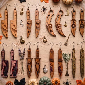 May include: A collection of wooden earrings shaped like knives, daggers, and cleavers, featuring engraved designs of skulls, spiderwebs, and stars. Some earrings have red paint accents, while others are plain wood. Small metal charms are scattered throughout.