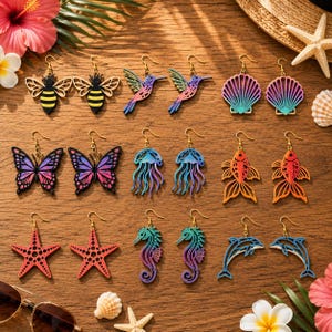 May include: A collection of nine pairs of dangle earrings featuring laser-cut designs including bees, hummingbirds, seashells, butterflies, jellyfish, goldfish, starfish, seahorses, and dolphins, all suspended from gold-toned metal earring hooks.