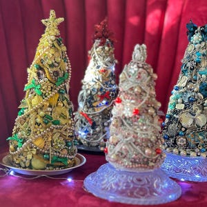 May include: Four decorative Christmas trees, each adorned with beads, jewels, and embellishments. The trees vary in color, including gold, silver, and blue, and are set on decorative bases. A gold star tops the first tree.
