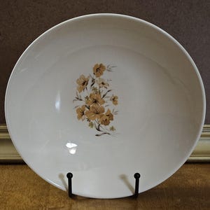 May include: Off-white vintage serving bowl with a floral design in shades of brown and yellow. The bowl is round and has a wide rim. The floral pattern is clustered on one side of the bowl. The bowl is displayed on a black metal stand.