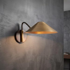 May include: A brass-colored wall sconce with a conical shade. The sconce has a dark, curved arm connecting the shade to a circular base. The fixture is mounted on a textured gray wall, casting a warm glow.