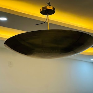 May include: A modern, bronze-colored pendant light fixture. The light has a large, shallow, bowl-shaped shade suspended from a gold-toned ceiling mount. The ceiling is white with recessed lighting and a yellow accent.