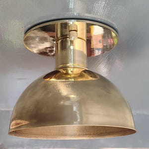 Puede incluir: Una lámpara en forma de cúpula de color latón. La lámpara tiene una base circular y un vástago cilíndrico, con una pantalla redondeada. El metal tiene un acabado pulido, que refleja el entorno. La lámpara está montada sobre una superficie gris.