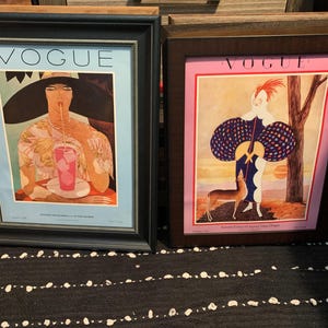 May include: Two framed Vogue magazine covers. The left frame has a black border and features a woman drinking a pink beverage. The right frame has a brown border and shows a woman with a deer. Both frames have the word "VOGUE" at the top.