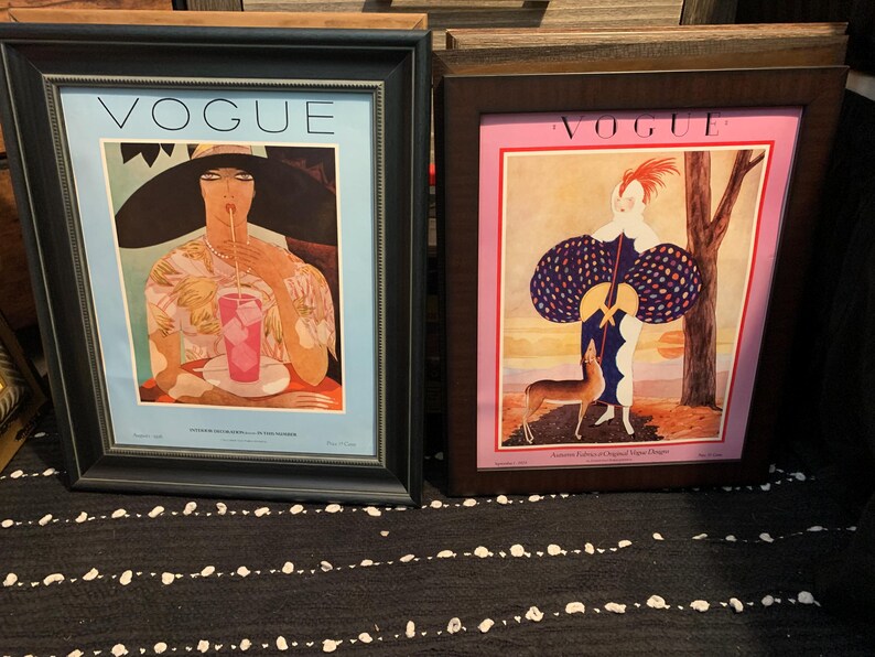May include: Two framed Vogue magazine covers. The left frame has a black border and features a woman drinking a pink beverage. The right frame has a brown border and shows a woman with a deer. Both frames have the word "VOGUE" at the top.