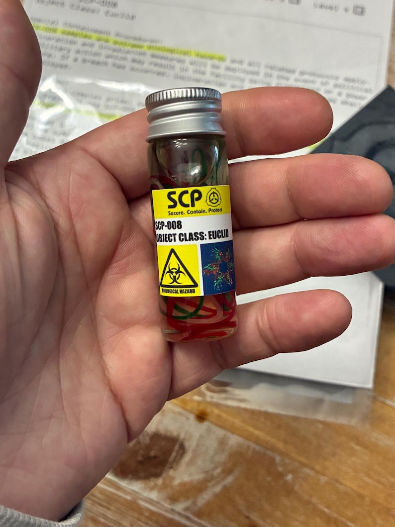 May include: A small clear glass vial with a silver screw-top lid containing red and green coiled filaments. A yellow and white label features the text SCP, SCP-008, OBJECT CLASS: EUCLID, and a yellow biological hazard warning symbol.