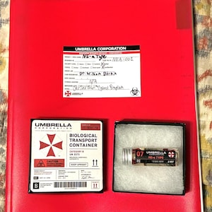 Puede incluir: Una carpeta roja brillante etiquetada como Umbrella Corporation con un documento de investigación, una caja de transporte biológico cuadrada y un pequeño vial de vidrio con tapa plateada en una caja negra, todo con la marca y texto de Umbrella Corporation.
