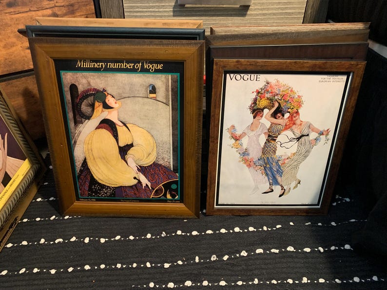 May include: Two framed vintage Vogue magazine covers. One features a woman in a yellow and purple dress with the text "Millinery number of Vogue". The other shows three women with floral headdresses.