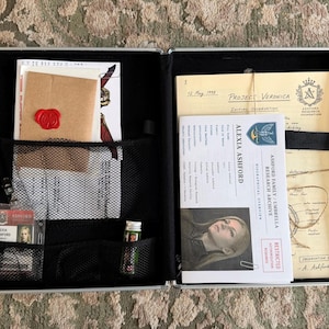 May include: A black metal briefcase containing documents and items including a brown envelope with a red wax seal, an Alexia Ashford identification badge, a small green vial, and papers labeled Project Veronica and Ashford Family Research Archive.