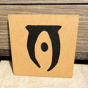 May include: Square cork board featuring a black symbol. The symbol is a stylized eye shape with a central dot. The cork board has a natural, light brown color and a textured surface.