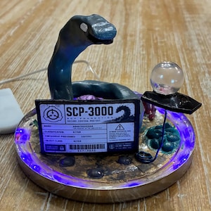 May include: A circular display base with purple LED lighting featuring a dark blue sculpted serpent figure. A rectangular sign reads SCP-3000 SCP FOUNDATION ANANTASHESHA. A small clear sphere sits on a black platform supported by a thin blue wire.