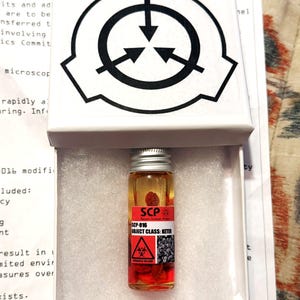 Peut inclure: Une petite fiole en verre remplie de liquide ambré et de particules rouges, avec une étiquette indiquant SCP-016 OBJECT CLASS: KETER et un symbole de risque biologique. La fiole est placée dans une boîte blanche avec le logo SCP noir sur le couvercle.