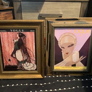 May include: Two framed vintage Vogue magazine covers. One features a woman in a long dress with the Vogue logo at the top. The other has a woman with a bird and the Vogue logo. Both are in gold frames.