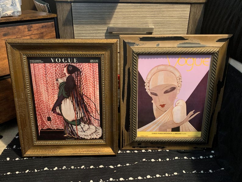 May include: Two framed vintage Vogue magazine covers. One features a woman in a long dress with the Vogue logo at the top. The other has a woman with a bird and the Vogue logo. Both are in gold frames.