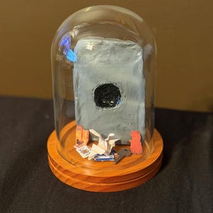 Peut inclure: Un dôme en verre transparent sur une base en bois contenant une dalle rectangulaire grise avec un trou circulaire noir au centre. À l'intérieur se trouvent une grue en papier blanc, une clé en métal, une carte à jouer et des découpages en papier.