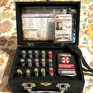 Peut inclure: Une boîte en bois noire à couvercle articulé, contenant des fioles de liquides colorés, une carte d'identité de risque biologique et une carte de la division des risques biologiques. La boîte a un fermoir en métal et le logo Umbrella Corporation est visible.