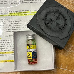 May include: A small clear glass vial with a silver screw cap containing liquid and red strands, featuring a yellow label with the text SCP-008, OBJECT CLASS: EUCLID, and a biohazard symbol. It is placed in a white box next to a black lid.