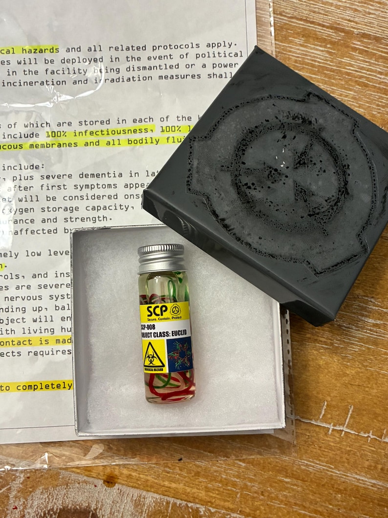 May include: A small clear glass vial with a silver screw cap containing liquid and red strands, featuring a yellow label with the text SCP-008, OBJECT CLASS: EUCLID, and a biohazard symbol. It is placed in a white box next to a black lid.