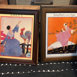 May include: Two framed vintage Vogue magazine covers. The left frame features a woman in a blue dress with a rooster, and the right frame shows a woman in a pink dress walking in a spring scene. Both frames have dark brown wooden frames.