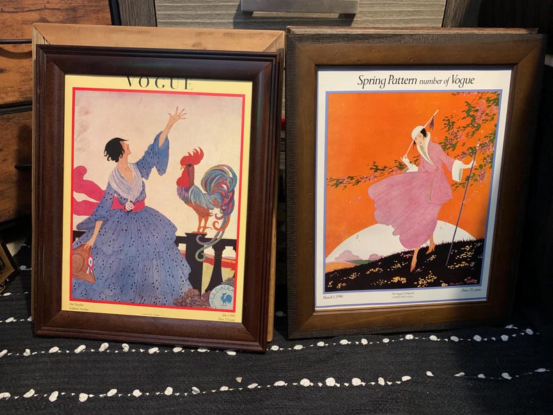 May include: Two framed vintage Vogue magazine covers. The left frame features a woman in a blue dress with a rooster, and the right frame shows a woman in a pink dress walking in a spring scene. Both frames have dark brown wooden frames.