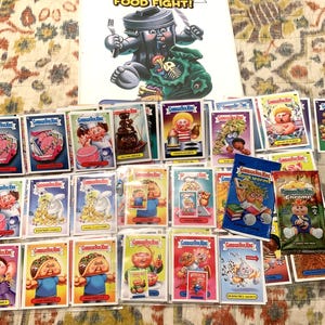 May include: A collection of Garbage Pail Kids trading cards and a promotional poster. The poster features a trash can character with a fork and knife. The cards have various cartoon characters and the text "Garbage Pail Kids".