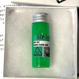 May include: A small clear glass vial filled with bright green liquid, topped with a silver screw cap. A label on the front reads SCP, SCP-447, OBJECT CLASS: SAFE, and includes a triangular warning symbol and a small circular image.