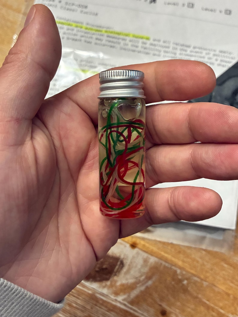 May include: A small, clear glass vial with a silver screw-top lid, filled with a transparent liquid and containing thin, tangled strands of red, green, and white material suspended inside the fluid, creating a colorful, abstract display.