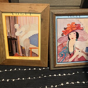 May include: Two framed vintage Vogue magazine covers. The left frame features a woman in a room, and the right frame showcases a woman in a large hat. Both frames have wooden borders, and the artwork is colorful.