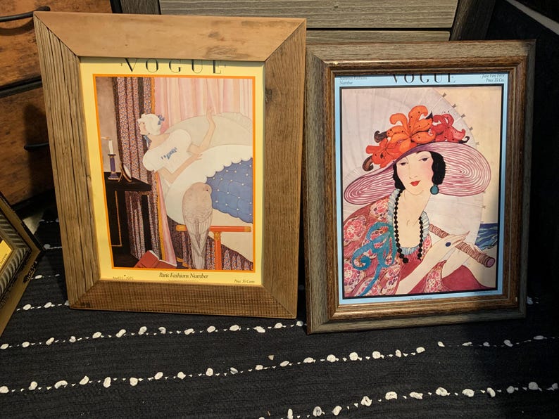 May include: Two framed vintage Vogue magazine covers. The left frame features a woman in a room, and the right frame showcases a woman in a large hat. Both frames have wooden borders, and the artwork is colorful.
