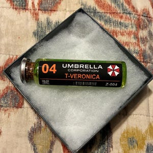 May include: A glass vial filled with a translucent green liquid, featuring a black label with the text 04, UMBRELLA CORPORATION, T-VERONICA, a red and white umbrella logo, a QR code, a barcode, and the code Z-004, resting in a square box.