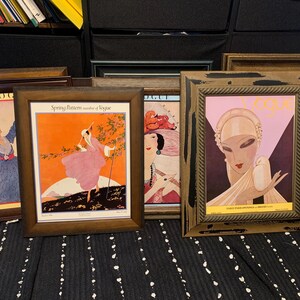 May include: A collection of framed vintage Vogue magazine covers. The frames are various shades of brown and gold. The artwork features illustrations of women in stylish attire, with vibrant colors and Art Deco designs. The covers are displayed on a black and white striped surface.