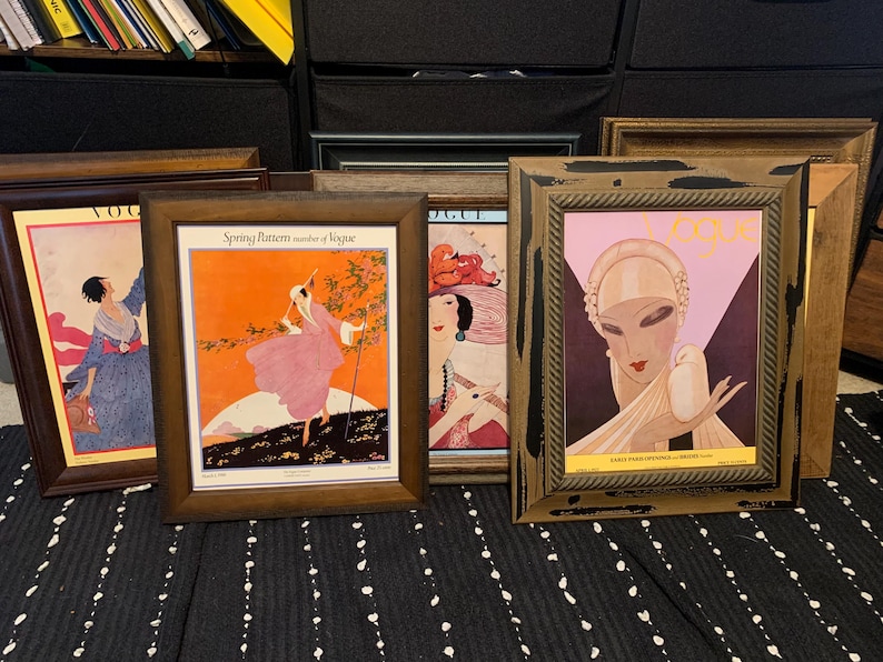 May include: A collection of framed vintage Vogue magazine covers. The frames are various shades of brown and gold. The artwork features illustrations of women in stylish attire, with vibrant colors and Art Deco designs. The covers are displayed on a black and white striped surface.
