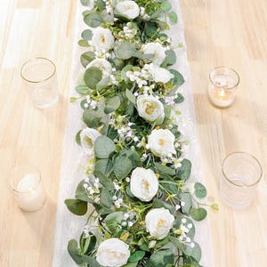 May include: A floral table runner featuring white roses and green eucalyptus leaves arranged on a white lace fabric strip. The arrangement is surrounded by four clear glass candle holders, with two containing lit white candles.