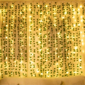 May include: A decorative curtain featuring multiple vertical strands of artificial green ivy leaves intertwined with warm white LED string lights. The lights create a soft glow against the foliage, hanging in a uniform pattern across the display.