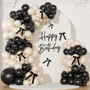 May include: A birthday party decoration featuring black and cream-colored balloons arranged in arches and clusters. Black ribbon accents are tied to the balloon clusters. The words "Happy Birthday" are displayed on a white backdrop.
