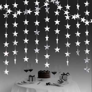 May include: A hanging decoration consisting of multiple vertical strings of silver star-shaped cutouts. The stars are arranged in a cascading pattern, suspended from a thin horizontal line against a dark background, creating a festive display.