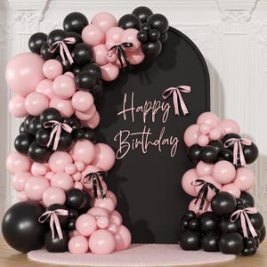 May include: A festive birthday backdrop featuring a black arch with the words "Happy Birthday" in cursive. The arch is adorned with clusters of black and pink balloons, some tied with pink ribbons. The scene is set on a pink rug.