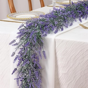 May include: A decorative garland featuring artificial lavender sprigs with vibrant purple flower clusters and green foliage. The long, flexible vine is arranged as a centerpiece, showcasing detailed botanical textures and realistic stems.