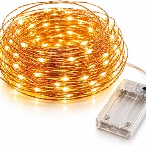 May include: A coiled strand of copper wire fairy lights emitting a warm yellow glow, connected to a clear rectangular plastic battery pack with a small black power switch and visible internal metal springs for battery placement.
