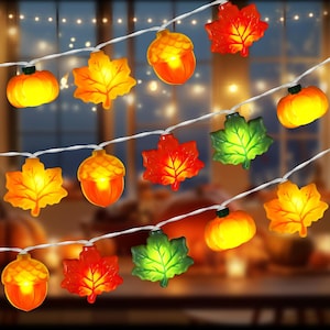 May include: A string of decorative lights featuring illuminated shapes of orange pumpkins, yellow and red maple leaves, and orange acorns. The translucent figures are connected by a thin white wire, creating a warm glow with autumn-themed motifs.