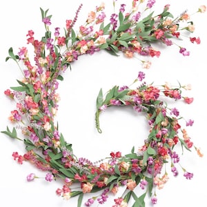 May include: A spiral-shaped floral garland featuring a mix of small pink, purple, and peach-colored blossoms interspersed with green leaves and slender lavender-like sprigs, arranged in a continuous, flexible vine structure on a plain background.