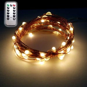 May include: A coiled string of copper wire fairy lights emitting a warm white glow, accompanied by a small rectangular remote control featuring buttons labeled with numbers one through eight, power symbols, and brightness adjustment icons.