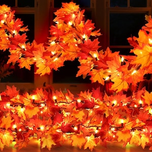 May include: A decorative garland featuring artificial maple leaves in vibrant shades of red, orange, and yellow. The foliage is intertwined with numerous small, warm-toned string lights that emit a soft glow throughout the dense leaf arrangement.