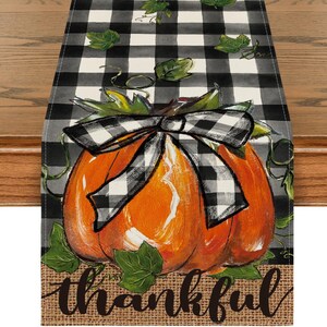 May include: A rectangular table runner featuring a black and white buffalo check pattern, a large orange pumpkin with green leaves, a black and white checkered bow tied around the pumpkin, and the word thankful written in black cursive script.