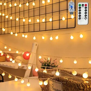 May include: A string of warm white LED globe lights featuring a USB power connector and a remote control with eight lighting modes and dimming functions. The lights are arranged in multiple strands, creating a glowing effect across the space.